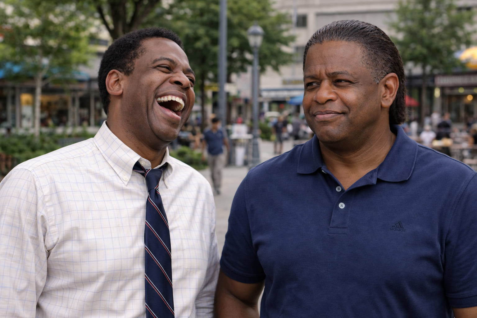 Two men outdoors sharing a broad laugh, one in black and one in a deep red shirt.